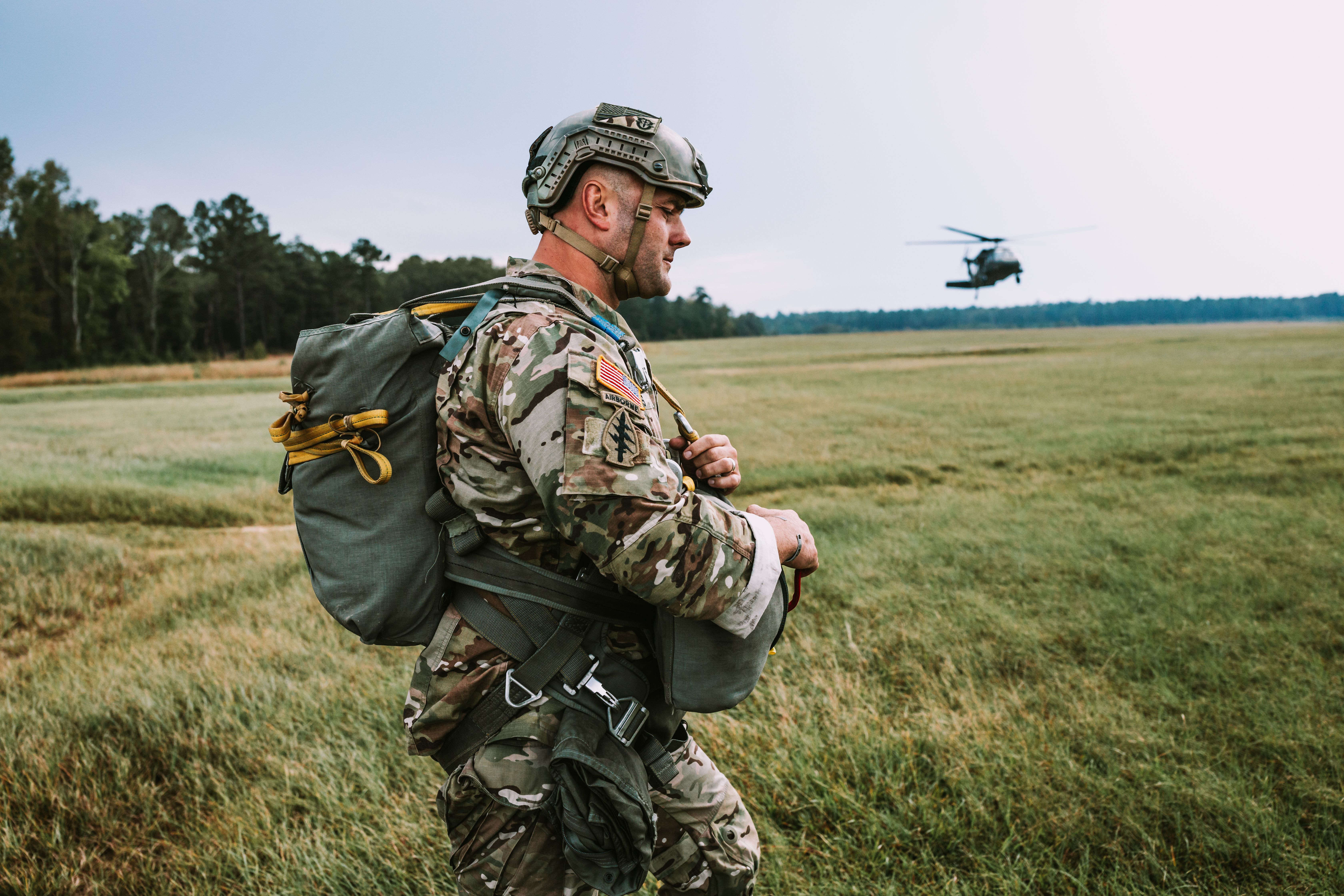 Soldier marching through low grassland during combined arms training with a helicopter in the background